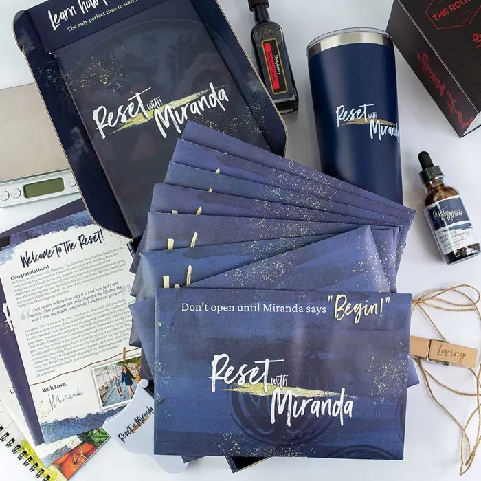 A Reset with Miranda box: a fanned set of dark blue sealed envelopes, a tumbler and a dropper bottle.